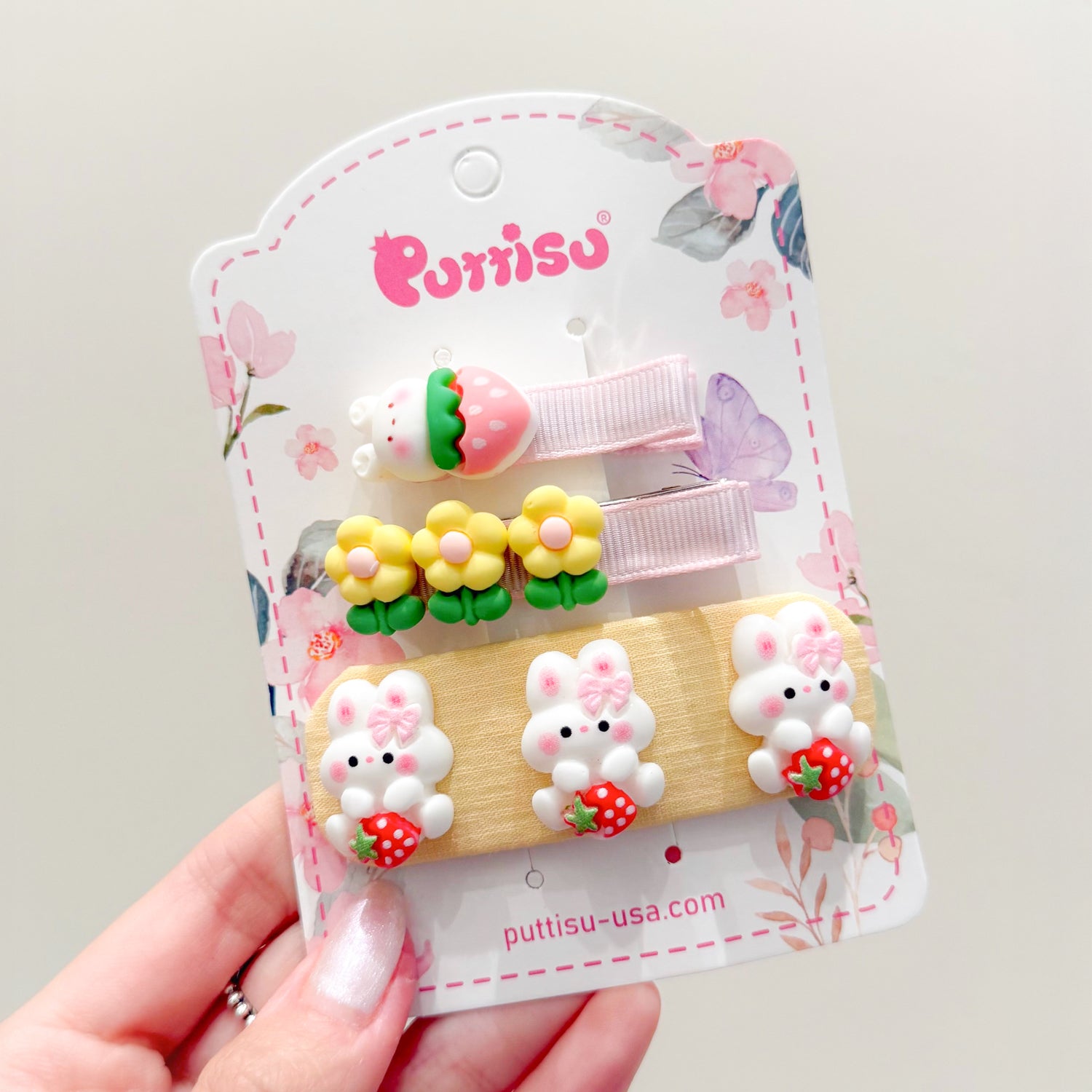 Three decorative hair clips on a floral-patterned card. The clips have strawberry-bear, flower, and bunny designs. A manicured hand holds the card.