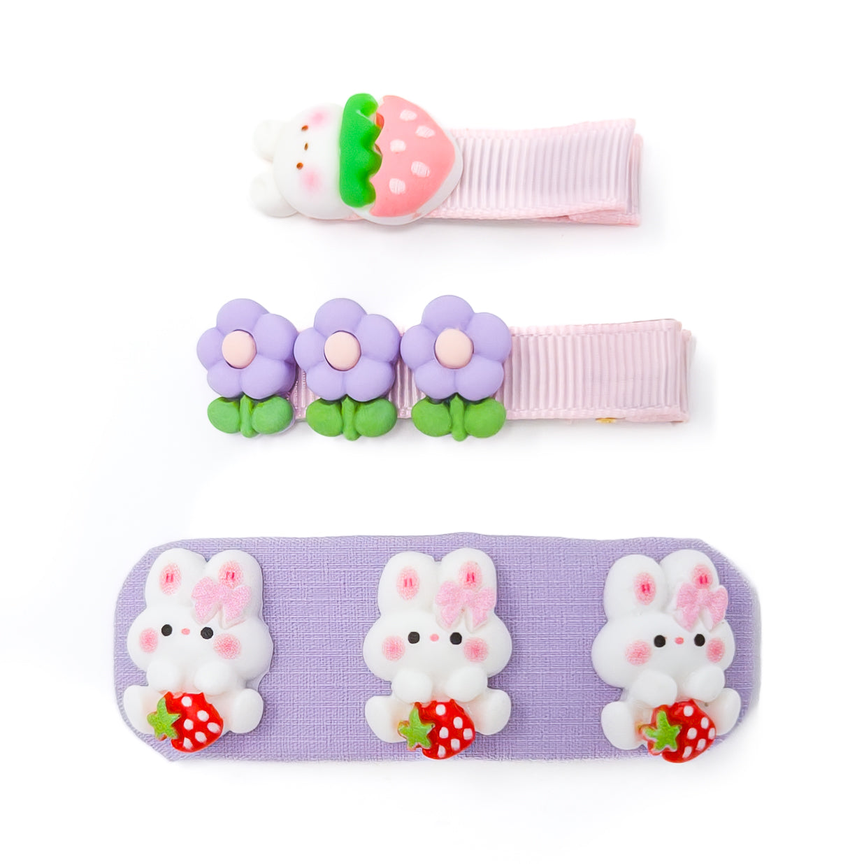 Three children's hair clips. One is light pink with a strawberry and bunny design. One is light pink with three purple flowers. The last one is purple and has three bunnies holding strawberries.