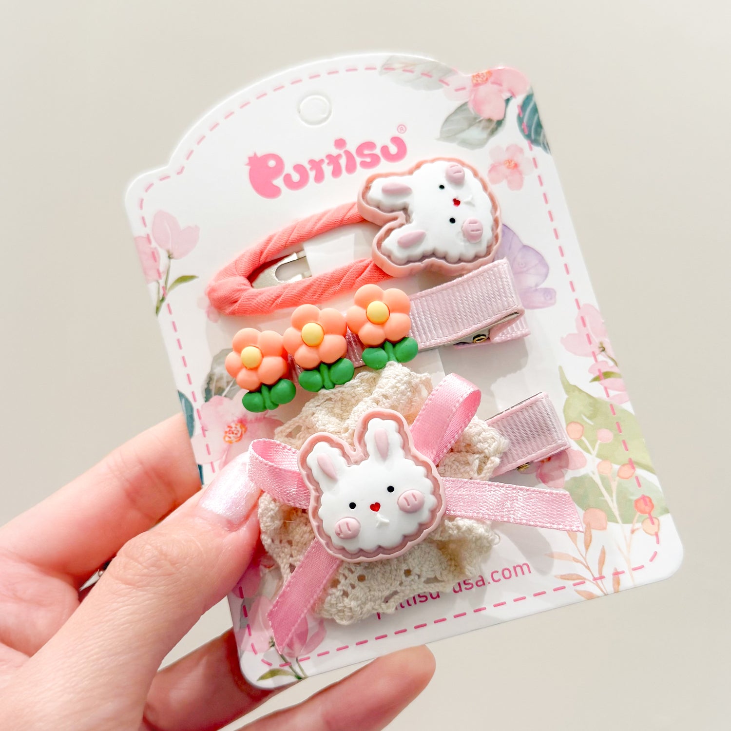 Close-up of a set of three children's hair clips on a floral card, featuring cartoon rabbits and flowers in shades of pink, peach, green, and white.