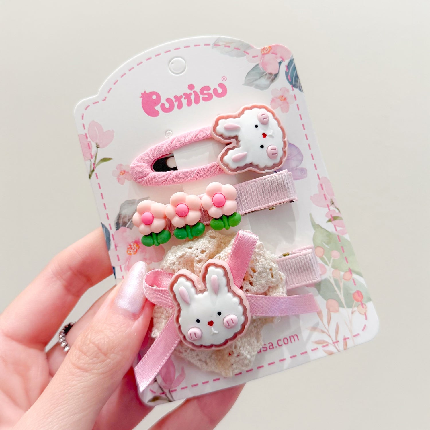 A hand holds a set of three children's hair clips on a floral-themed card, featuring a bunny, flowers, and pink ribbons.