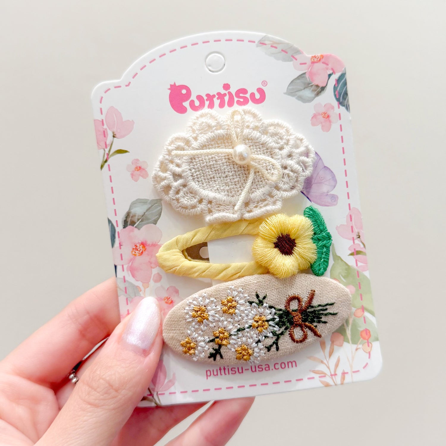 A hand holds a card of three hair clips with embroidered floral designs.