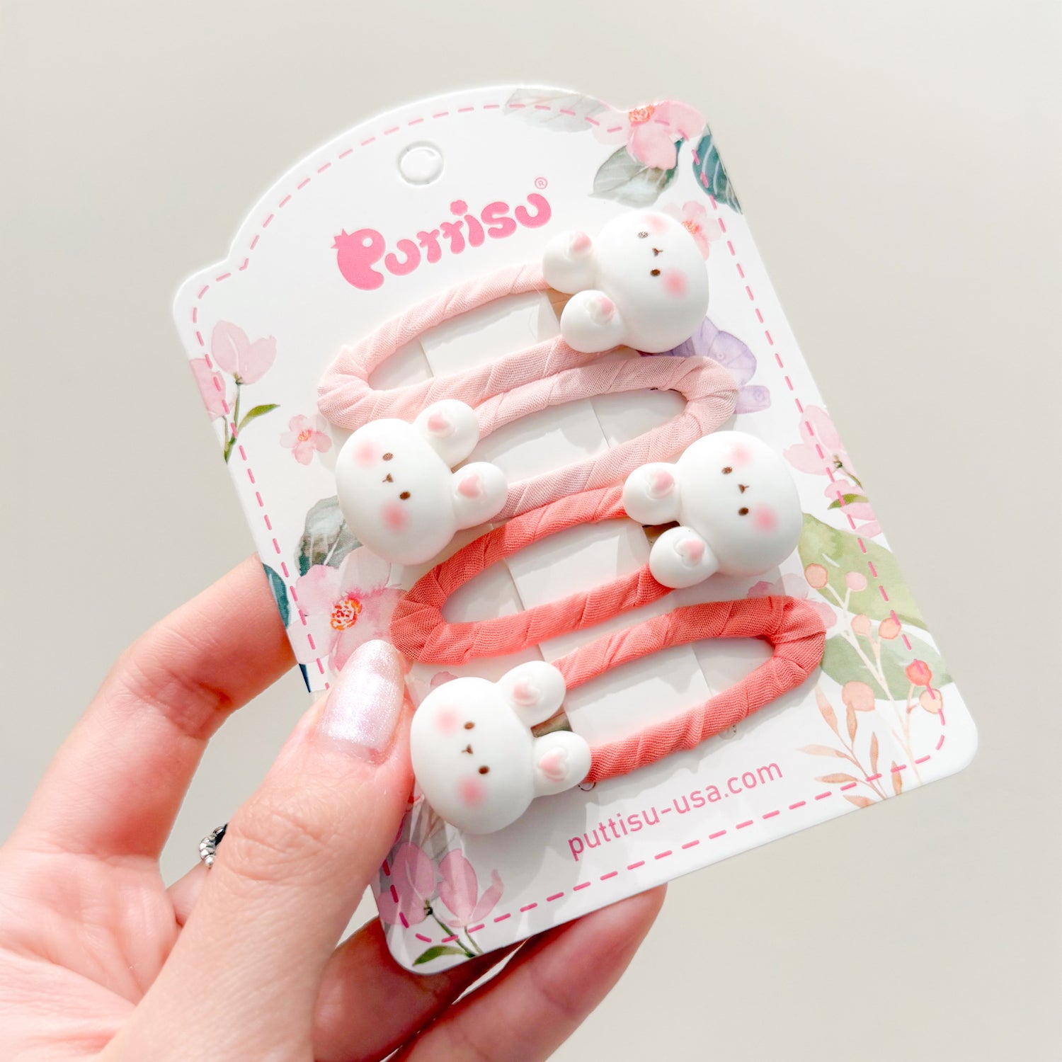 Three hair clips with small white bunny faces on pink and orange fabric strips, displayed on a floral-themed product card held in a hand.
