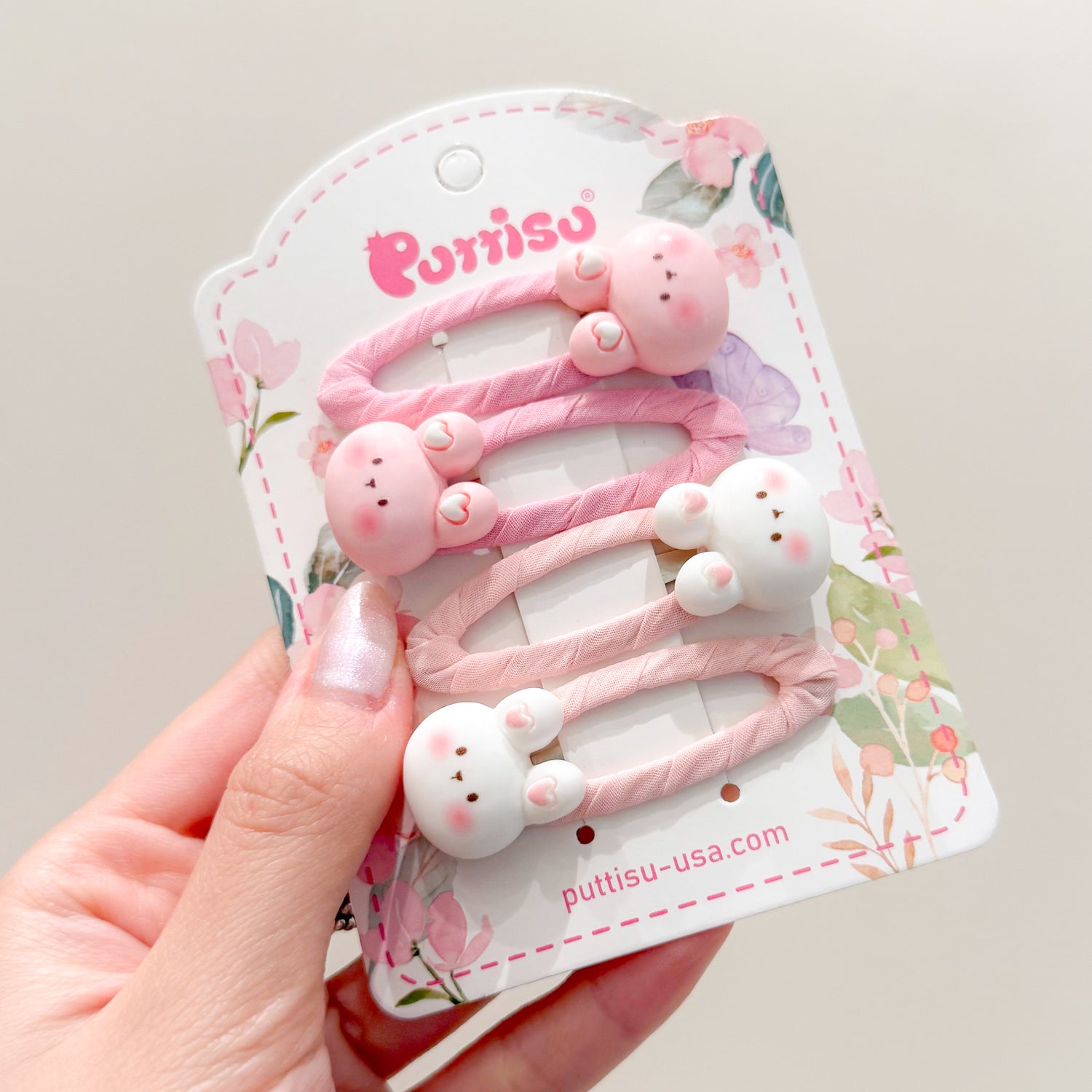 A hand holding a product display card with three pink and white bunny-shaped hair clips.