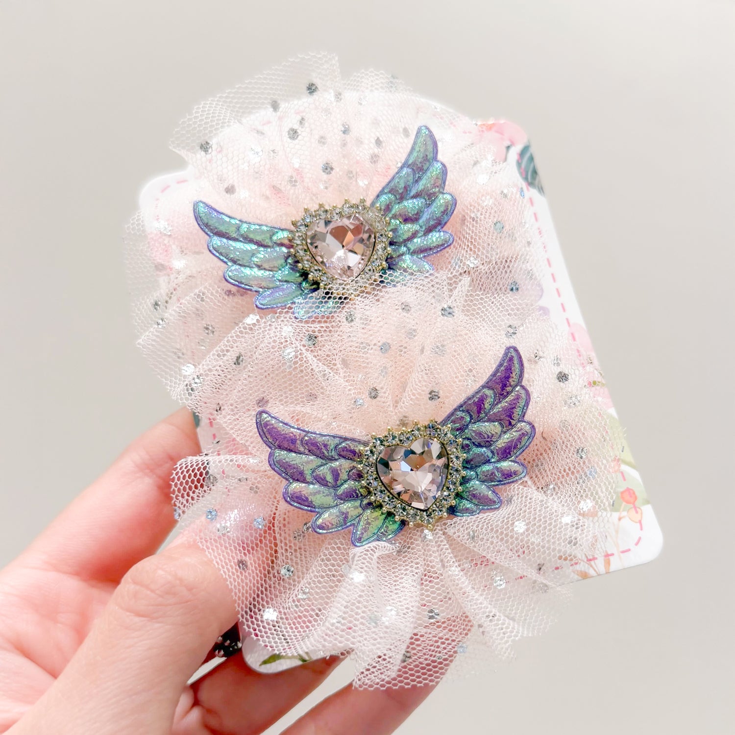 Close-up of two decorative hair clips with iridescent wings and heart-shaped jewels, on a white, glittery mesh backing, held in a hand.
