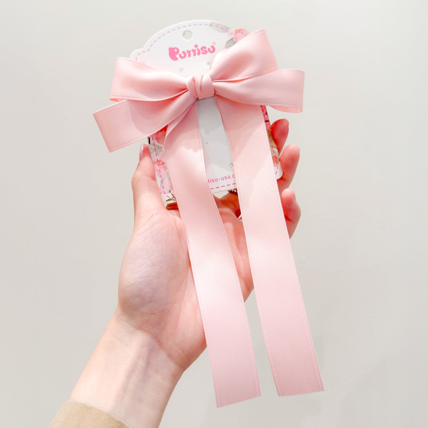 Hand holding a light pink satin ribbon hair clip with a bow and trailing ribbons on a floral-patterned tag.