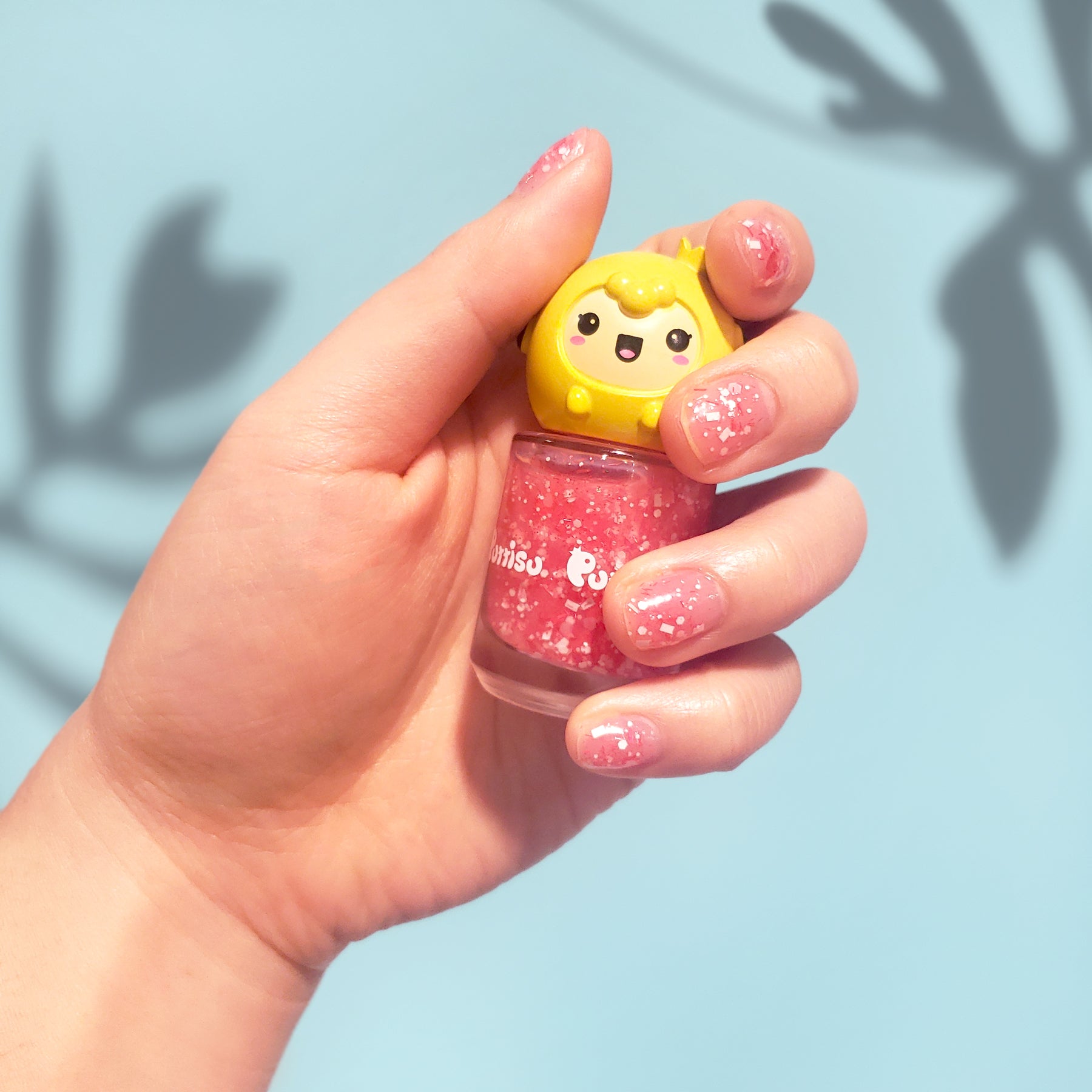 Hand holding pink glitter nail polish bottle with a yellow cartoon character cap.