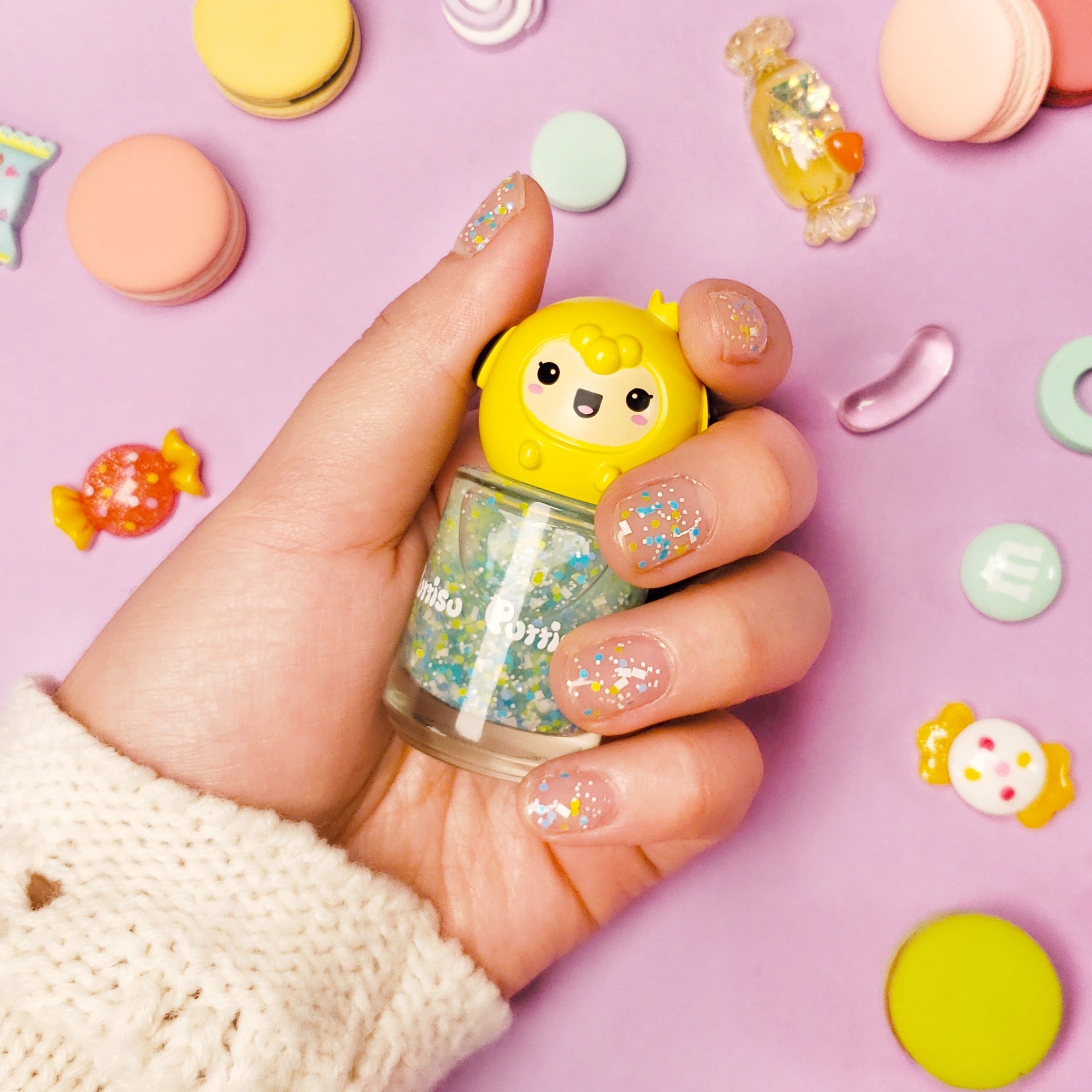 Hand holding a bottle of nail polish with a cartoon character cap, surrounded by pastel-colored candies and macarons, over a lavender surface.