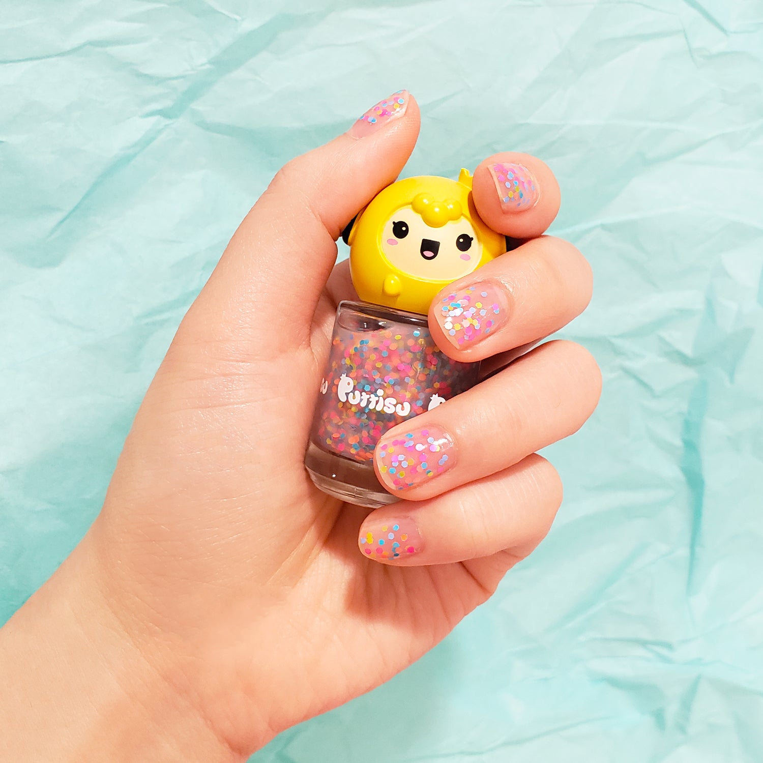 A hand holding a bottle of Purrissu nail polish with a yellow, cartoon-like cap, and matching glitter nail polish on the fingernails, against a turquoise background.