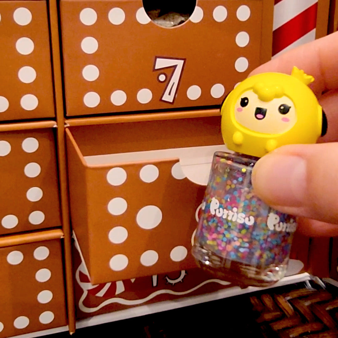 Close-up of an advent calendar with polka dots and drawer number 7 open, revealing a bottle of glitter nail polish with a cartoon chick on top held by a hand.