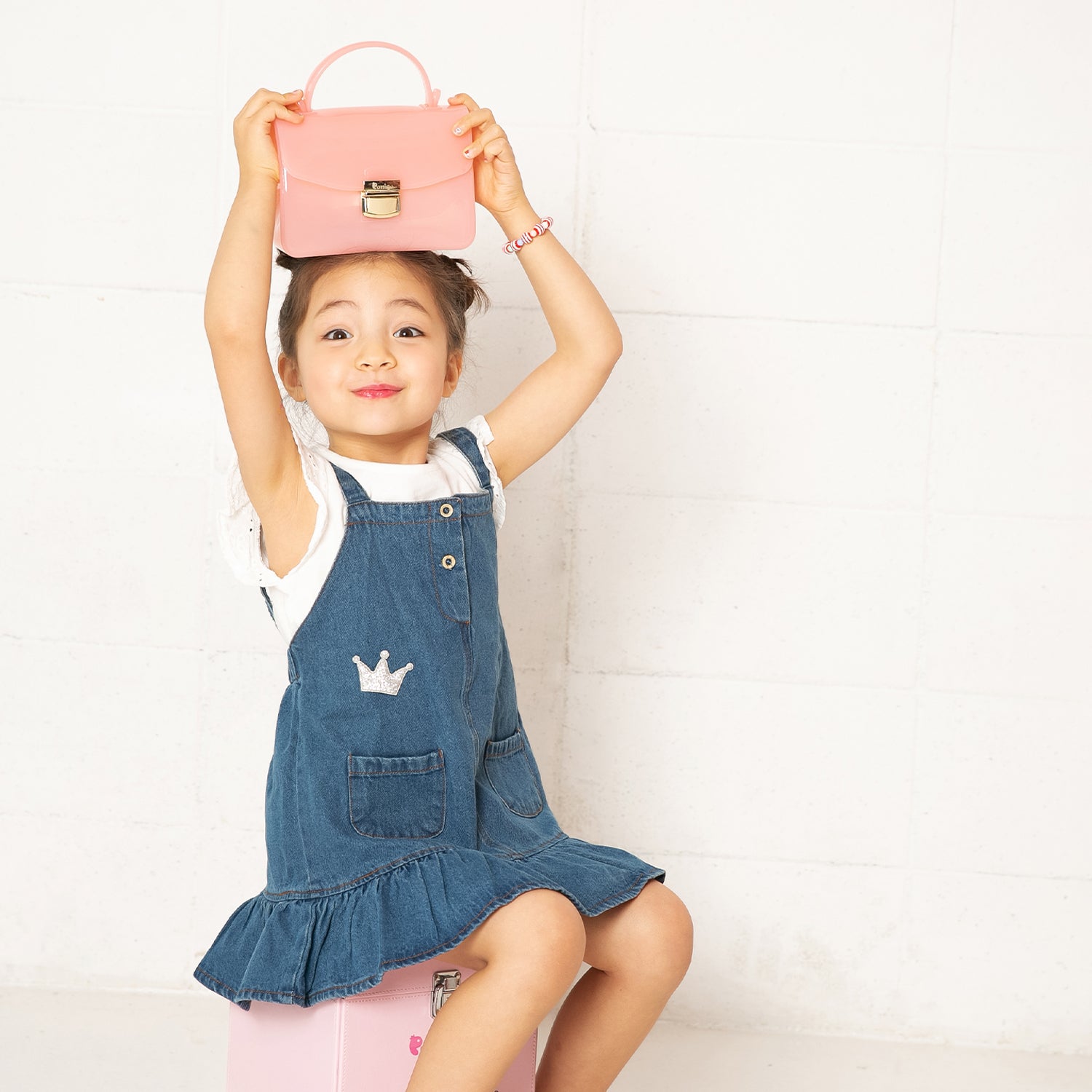 Young girl sitting with a pink purse atop her head, wearing a denim dress with a crown embellishment.