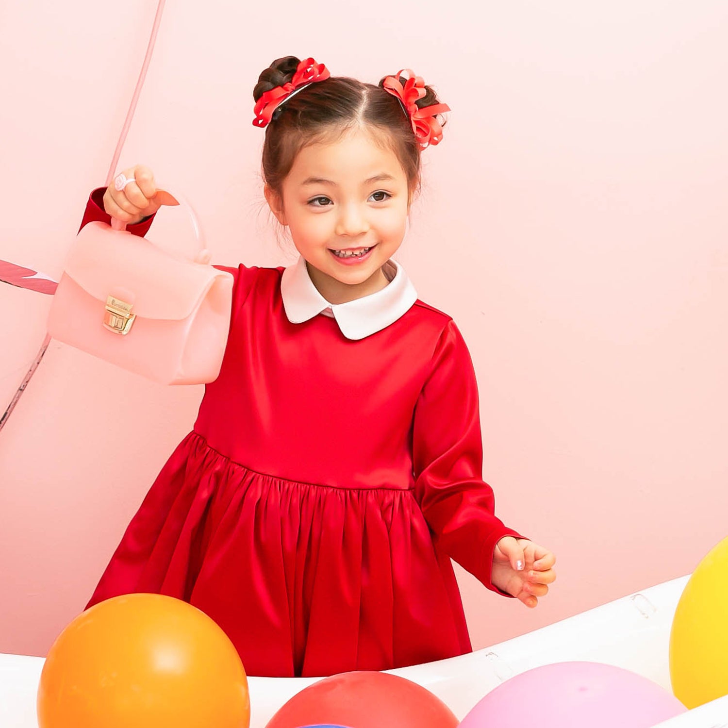 Young girl in red dress with white collar holding a pink purse, surrounded by colorful balloons.