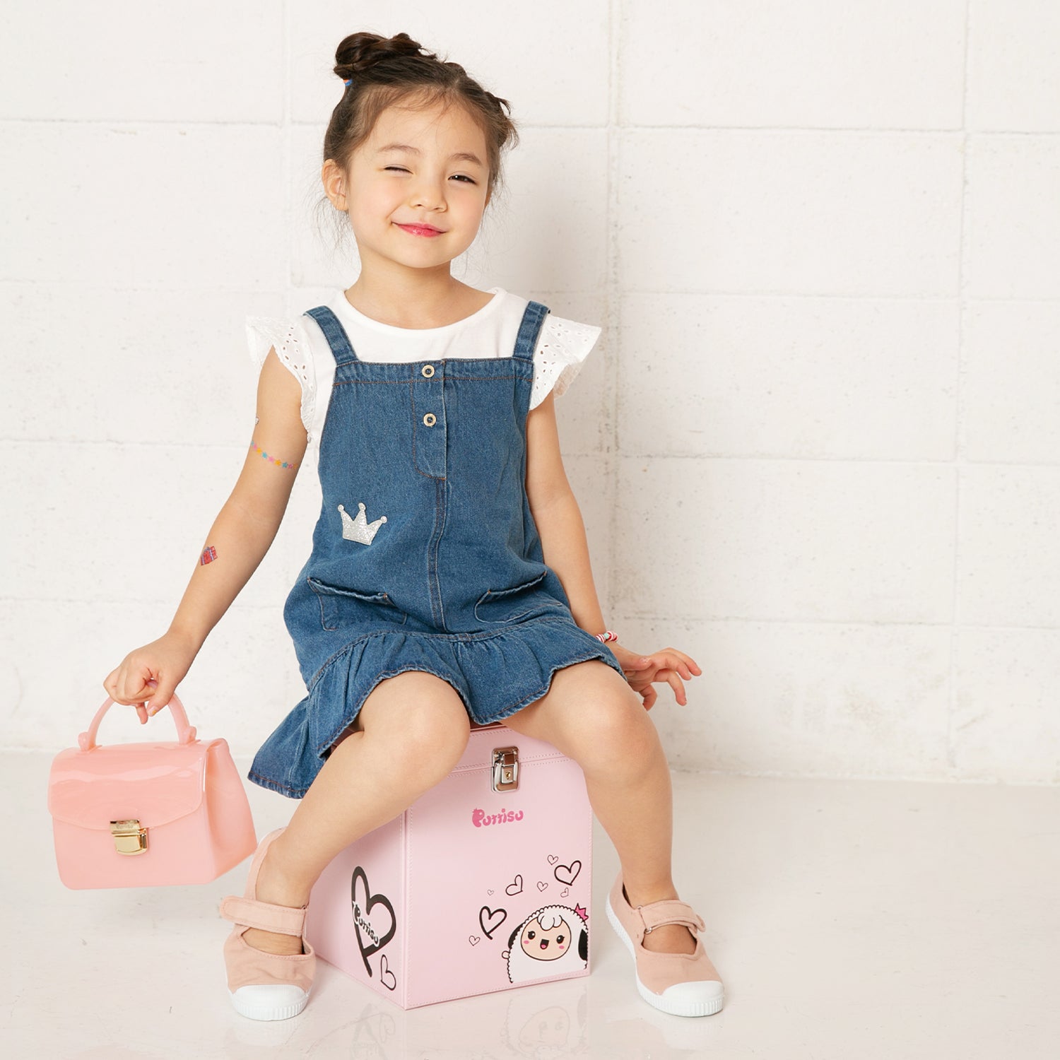 Girl in denim dress with pink accessories sitting on a pink box.