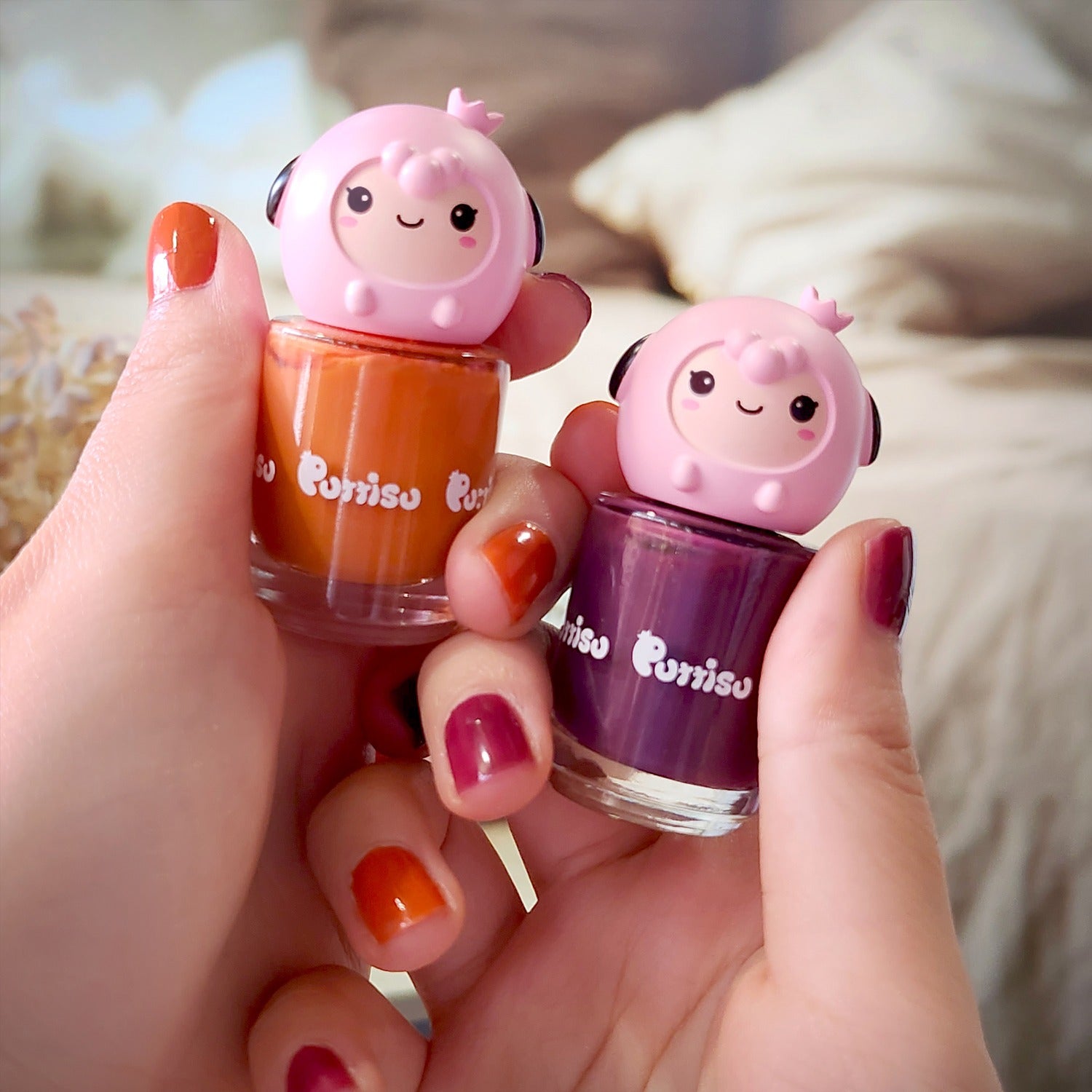 Two hands holding nail polish bottles. The left hand holds an orange bottle; the right hand holds a purple bottle. The bottle tops are pink cartoon sheep. The nails are painted with colors that match the bottles.