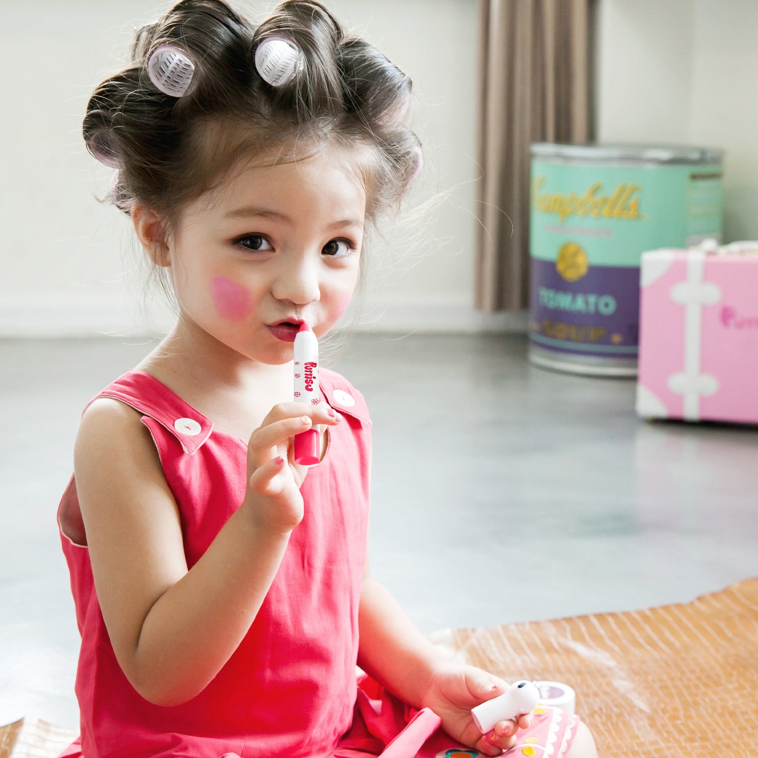 A young girl with hair rollers, wearing a red dress and applying lipstick.