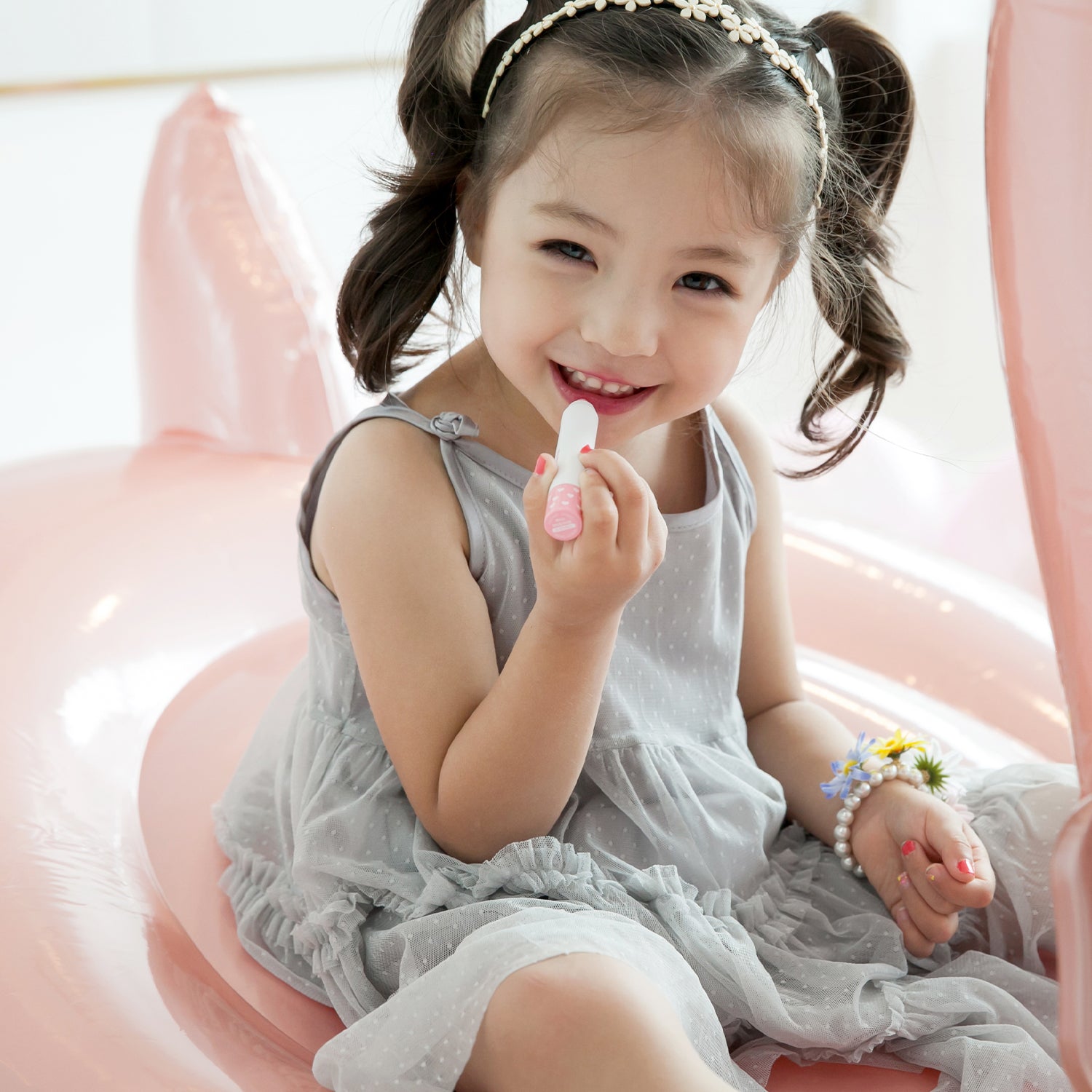 Little girl with pigtails and a flower headband applying lipstick while sitting on a pink inflatable swan. She wears a grey, polka-dotted dress and a pearl bracelet with flowers.