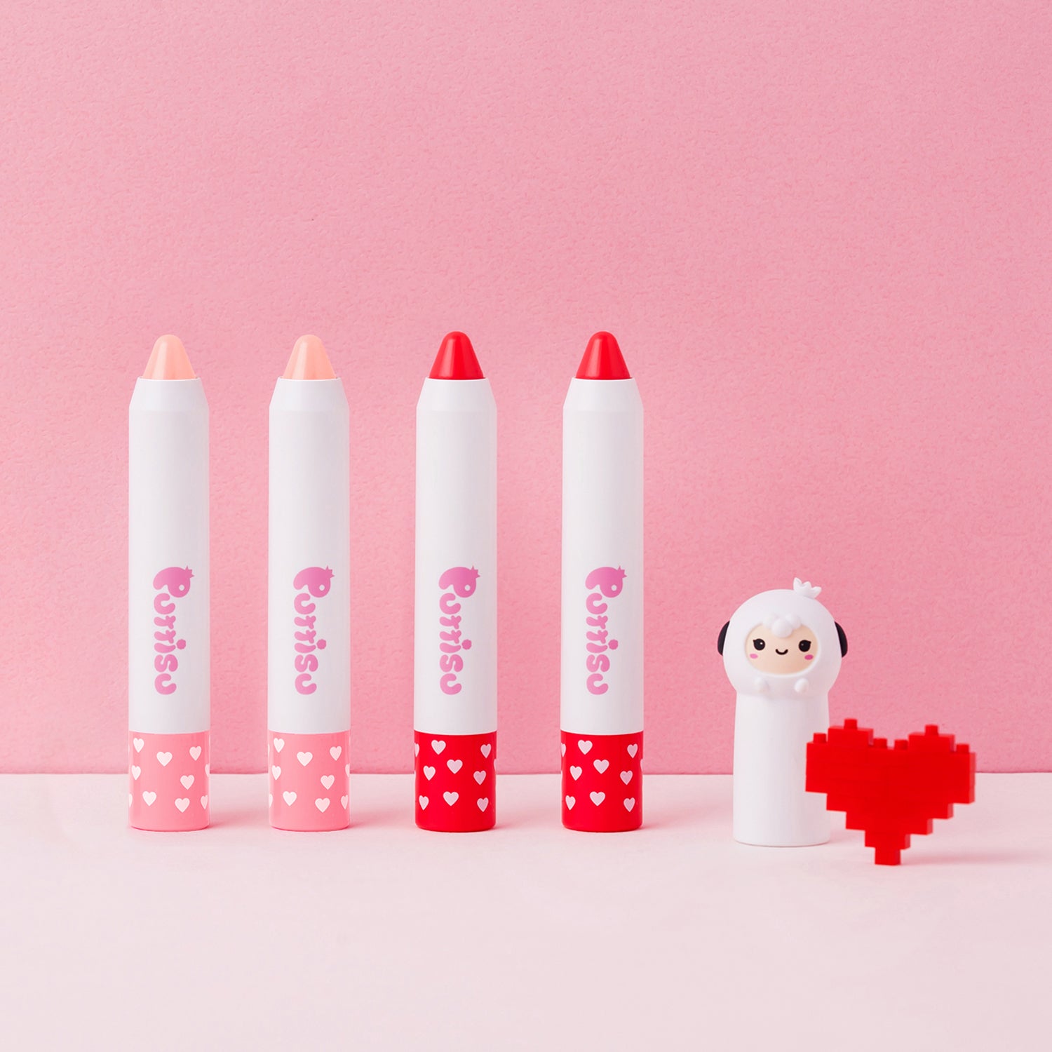 Four lip crayons, a sheep figurine, and a block heart on a pink surface.