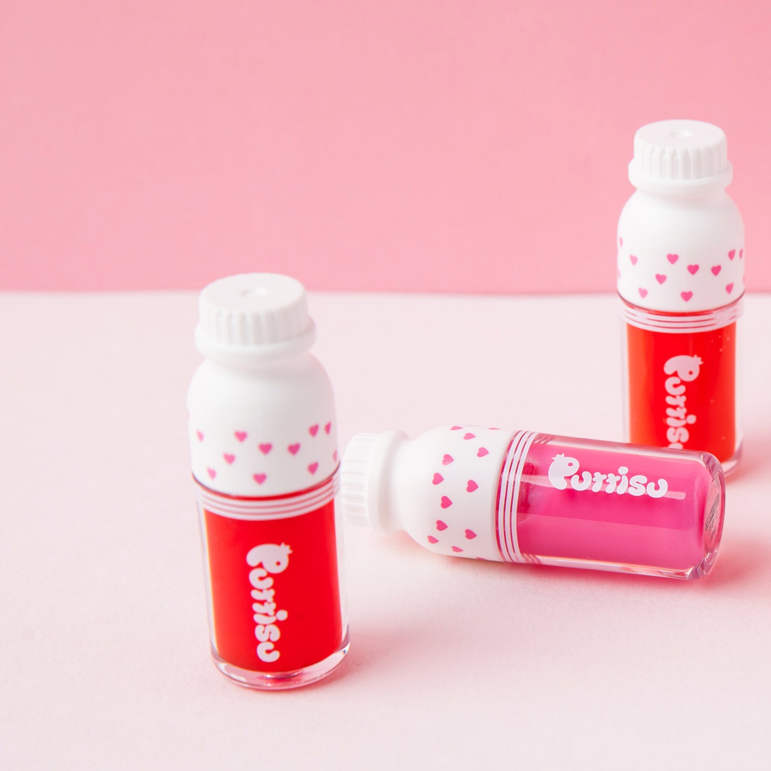 Three small, milk-bottle-shaped containers of "Purrisu" lip tint, one red, one pink, and one standing upright, against a pink background.