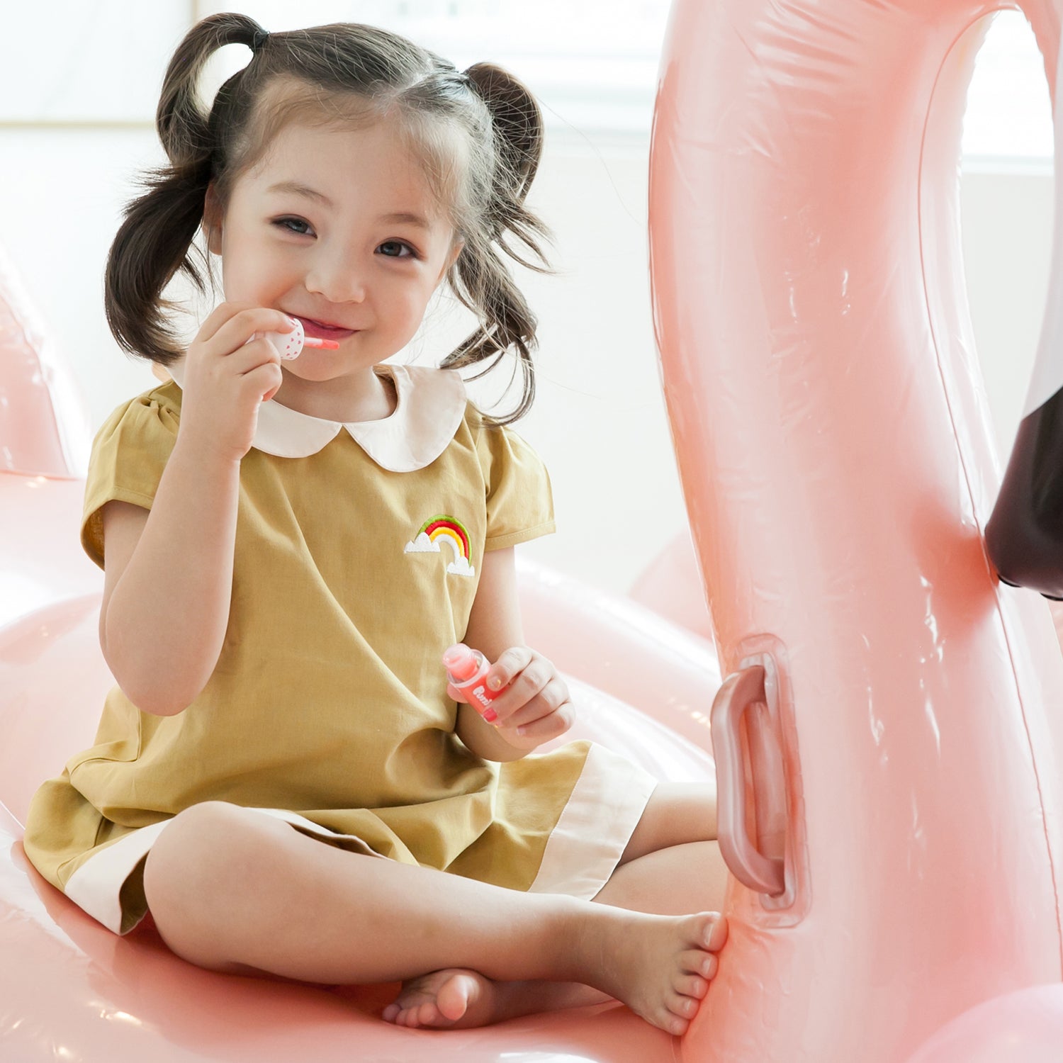 Young girl in yellow dress with rainbow patch, sitting on pink inflatable swan with lipstick.