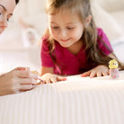 A woman paints a young girl's nails with pink nail polish on a white bed. A yellow and white nail polish bottle sits to the right.