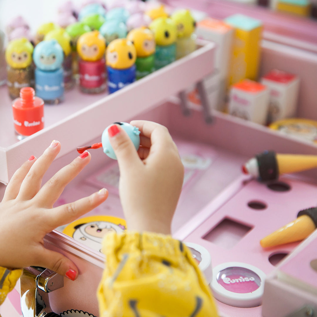 Close-up of a child's hands applying red nail polish from a light blue bottle to their nails, surrounded by a pink play makeup kit containing colorful character-themed nail polish bottles, toy ice cream cones, and blush compacts, against a soft, blurred background.