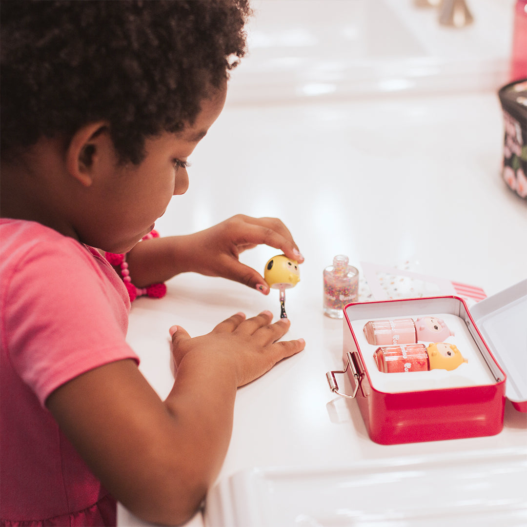 A young child in a pink shirt applies nail polish at a white counter with a red tin box of additional nail polish.