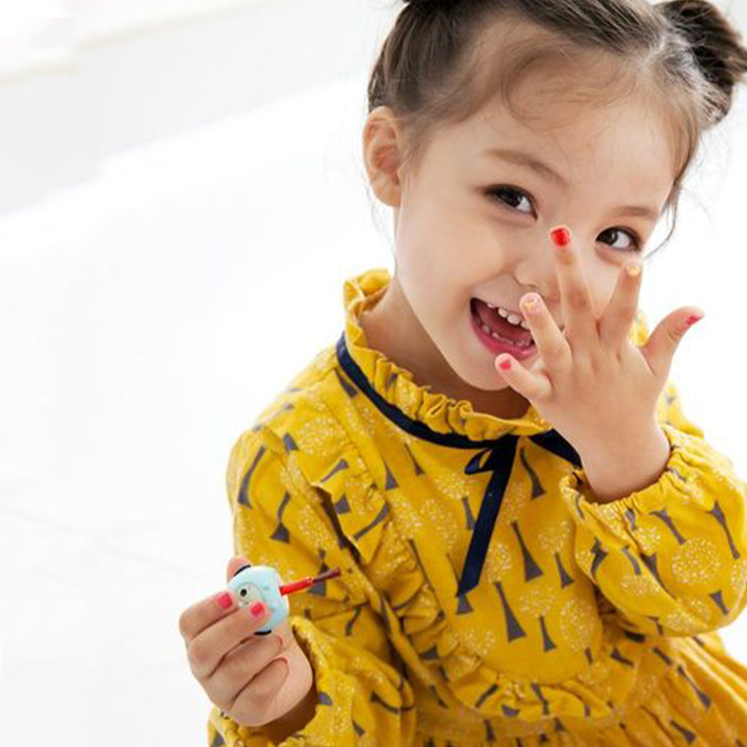 A little girl in a yellow dress with a navy blue bow and accents holds up her fingers showing painted nails and a blue nail polish bottle.