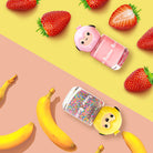 Overhead shot of pink nail polish with a sheep head and iridescent glitter polish with a chick head, with strawberries on a yellow background and bananas on a pink background.