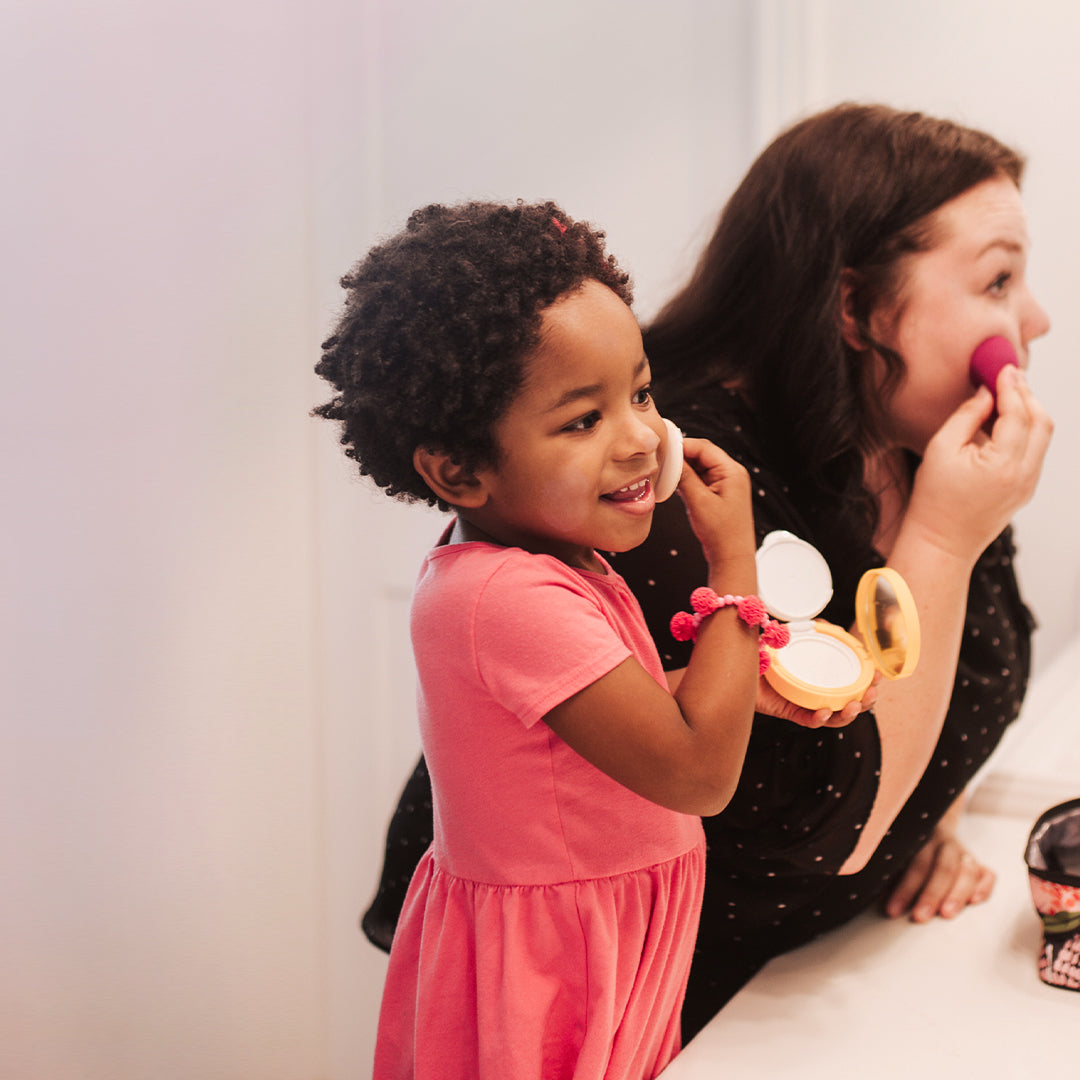 A little girl in a pink dress plays with makeup alongside her mother in the bathroom.