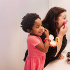 A little girl in a pink dress plays with makeup alongside her mother in the bathroom.