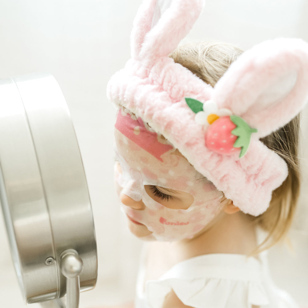 kid wearing a pink plush robe and bunny ear plush spa headband with a Puttisu face sheet mask against a white background