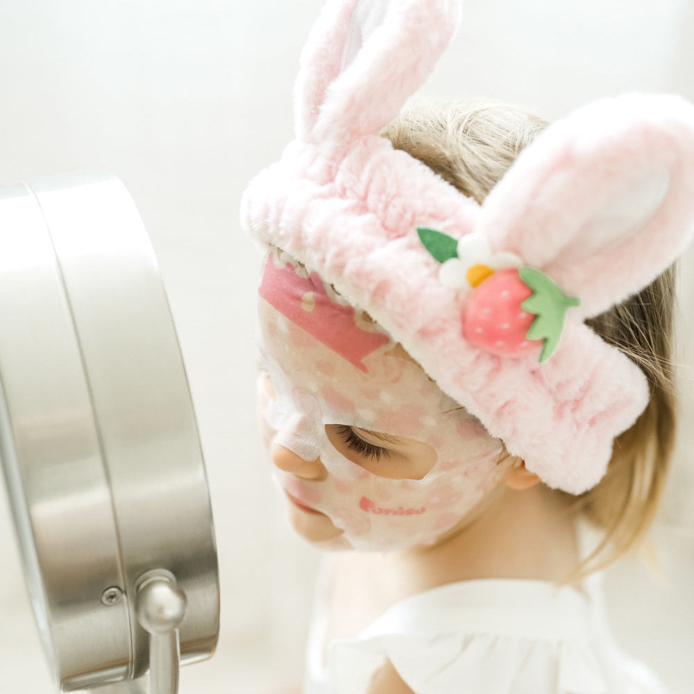 kid wearing a pink plush robe and bunny ear plush spa headband with a Puttisu face sheet mask against a white background