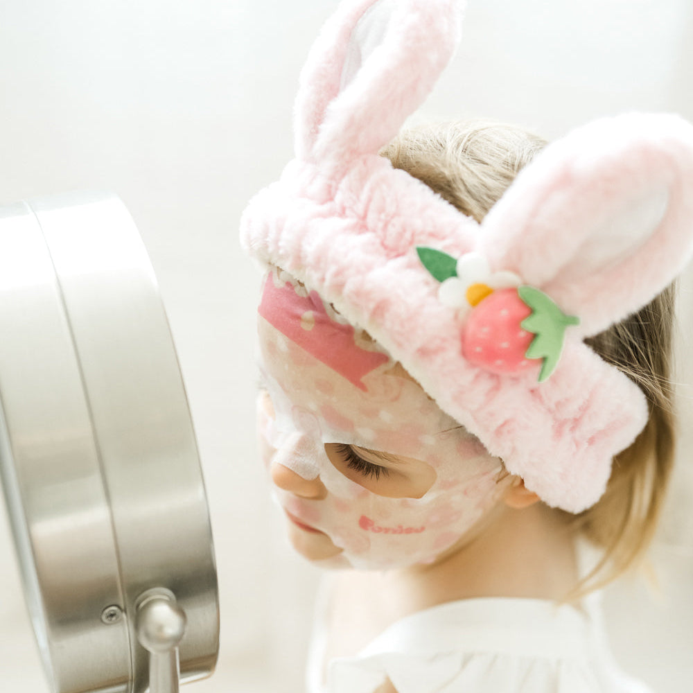 kid wearing a pink plush robe and bunny ear plush spa headband with a Puttisu face sheet mask against a white background