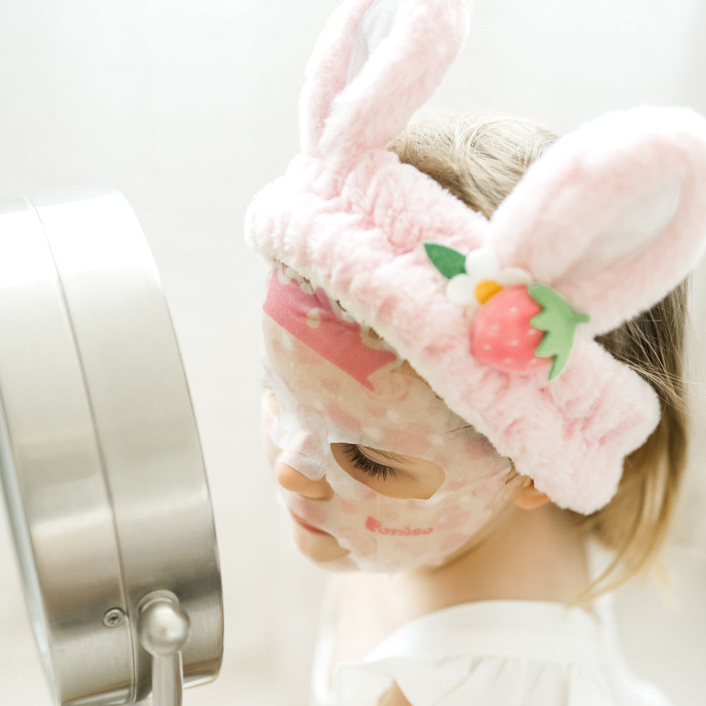 kid wearing a pink plush robe and bunny ear plush spa headband with a Puttisu face sheet mask against a white background