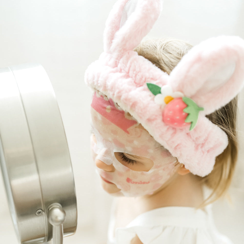 kid wearing a pink plush robe and bunny ear plush spa headband with a Puttisu face sheet mask against a white background