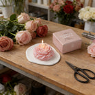 Pink peony flower soy wax candle by Jellycup, on a white ceramic dish near some roses and a candle box.