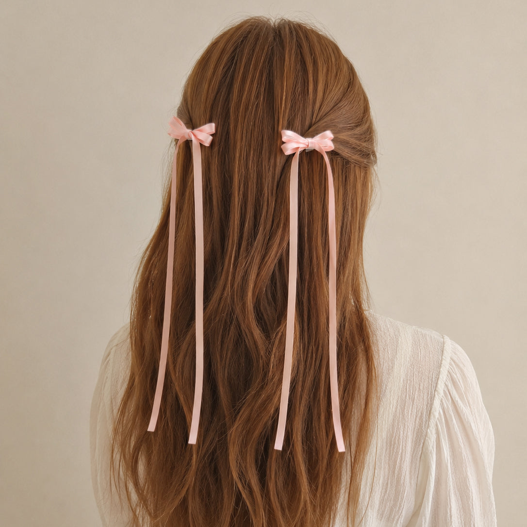 Pink ribbon bow hair clips with long flowing ribbons, styled on wavy brown hair from the back.
