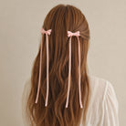 Pink ribbon bow hair clips with long flowing ribbons, styled on wavy brown hair from the back.
