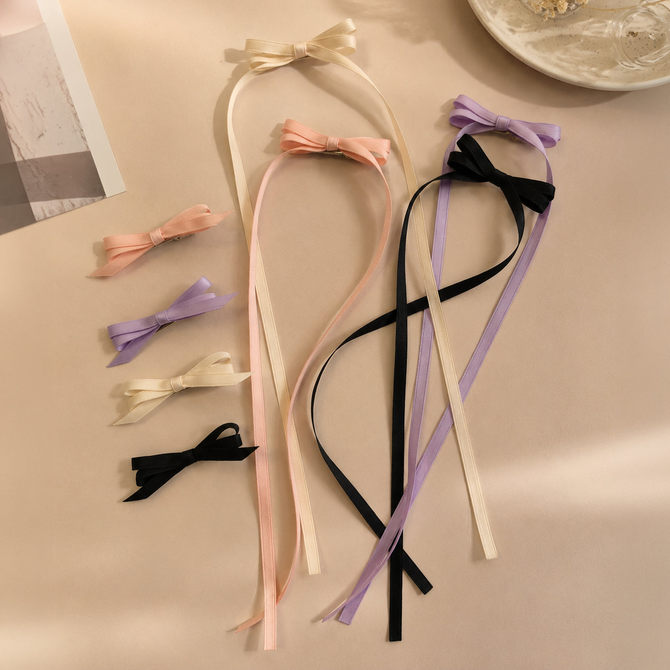 Four ivory ribbon bow clips are displayed among pink, lavender, and black ribbon bow clips, with long ribbon streamers, on a light surface.