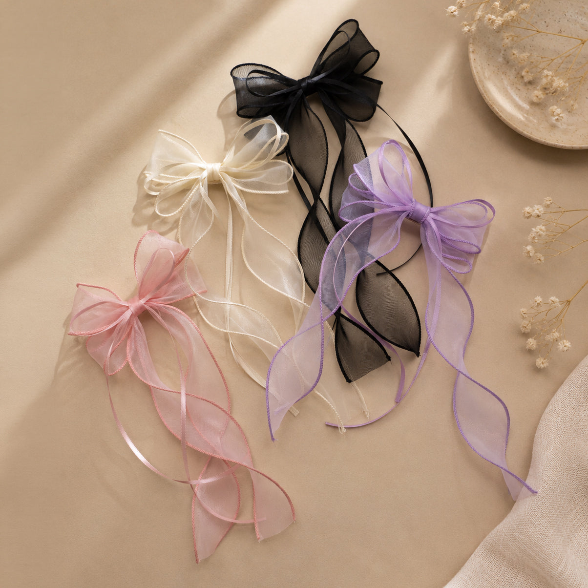 Four sheer organza double ribbon clips in black, cream, pink, and purple, with long, flowing tails on a beige surface.