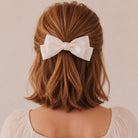 Ivory large satin double bow clip in wavy reddish-brown hair above a light blouse, seen from the back.