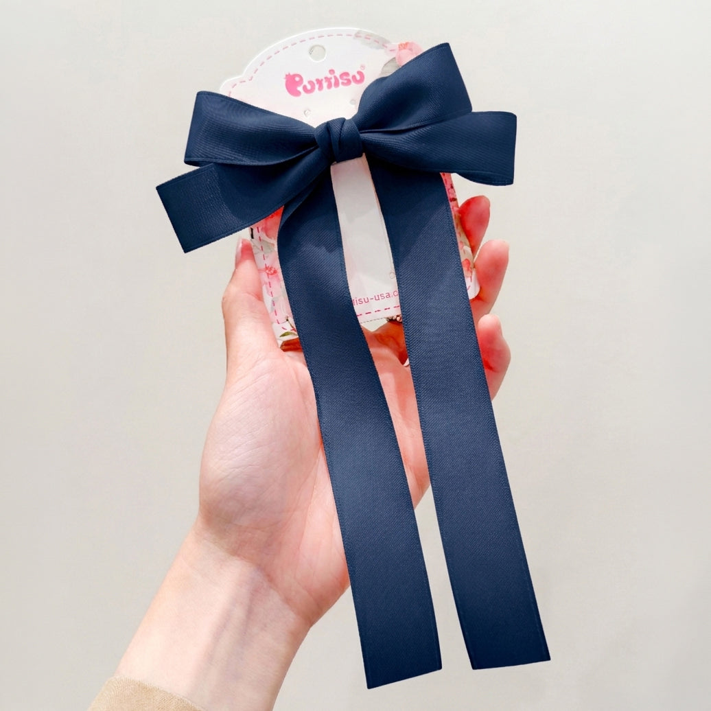 Hand holding a navy satin ribbon hair clip with a bow and trailing ribbons on a floral-patterned tag.
